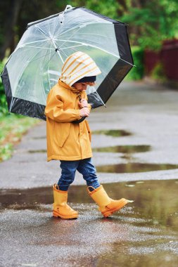 Kid in yellow waterproof cloak, boots and with umbrella playing outdoors after the rain.