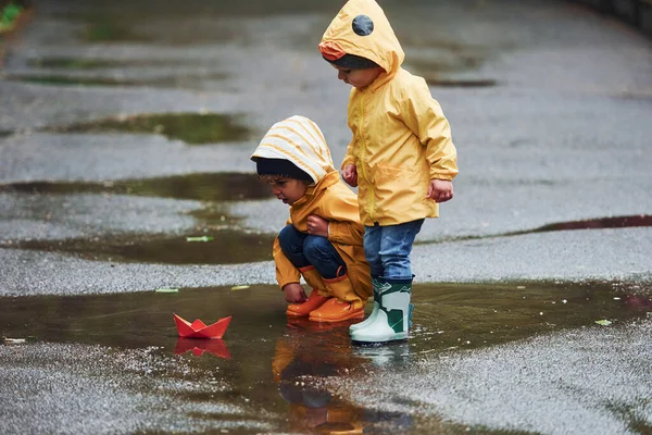 Two Kids Yellow Waterproof Cloaks Boots Playing Handmade Paper Boat — Zdjęcie stockowe