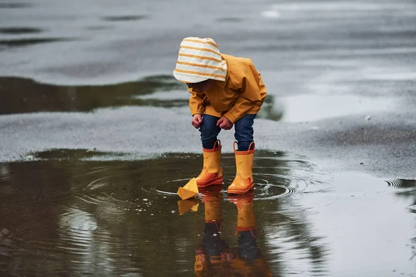 Sarı su geçirmez pelerinli ve botlu çocuk yağmurdan sonra el yapımı tekne oyuncağıyla oynuyor..