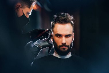 Siyah koruyucu maskeli bir adam berber dükkanında oturup saçlarını kestiriyor..