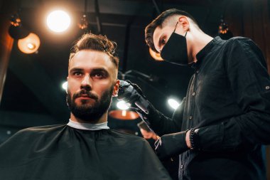 Siyah koruyucu maskeli bir adam berber dükkanında oturup saçlarını kestiriyor..