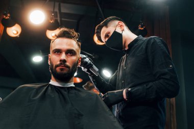 Siyah koruyucu maskeli bir adam berber dükkanında oturup saçlarını kestiriyor..