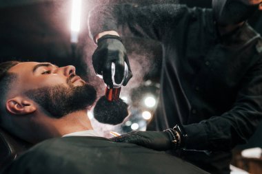 Şık bir saç stili olan genç adam berber dükkanında sakalını kestiriyor..