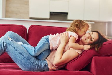 Lying down on red sofa. Young mother with her little daughter in casual clothes together indoors at home.