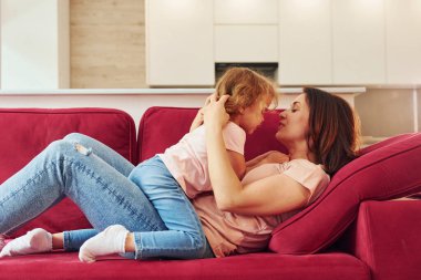 Lying down on red sofa. Young mother with her little daughter in casual clothes together indoors at home.