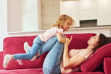 Lying down on red sofa. Young mother with her little daughter in casual clothes together indoors at home.