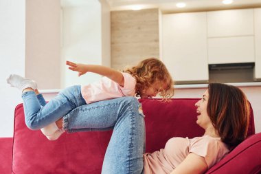 Having fun on red sofa. Young mother with her little daughter in casual clothes together indoors at home.