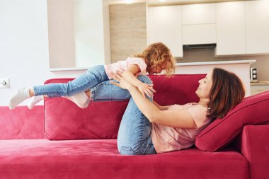 Having fun on red sofa. Young mother with her little daughter in casual clothes together indoors at home.