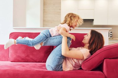Having fun on red sofa. Young mother with her little daughter in casual clothes together indoors at home.