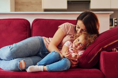 Lying down on red sofa. Young mother with her little daughter in casual clothes together indoors at home.