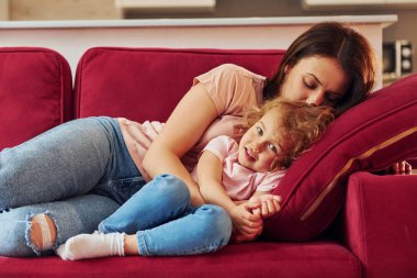 Lying down on red sofa. Young mother with her little daughter in casual clothes together indoors at home.