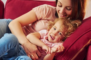 Lying down on red sofa. Young mother with her little daughter in casual clothes together indoors at home.