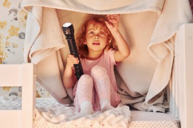 El feneriyle battaniyenin altında oturmak. Gündüz vakti evde rahat giyimli şirin küçük bir kız var..