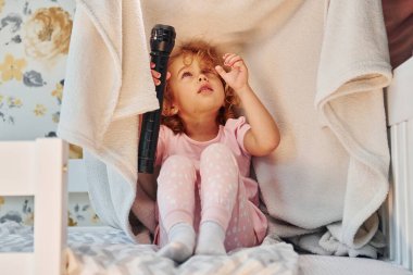 El feneriyle battaniyenin altında oturmak. Gündüz vakti evde rahat giyimli şirin küçük bir kız var..