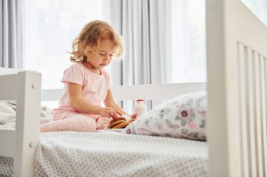 Yatakta oturmak, kurabiye yemek ve süt içmek. Gündüz vakti evde rahat giyimli şirin küçük bir kız var..