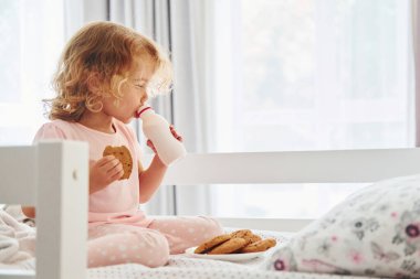Yatakta oturmak, kurabiye yemek ve süt içmek. Gündüz vakti evde rahat giyimli şirin küçük bir kız var..