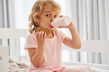 Yatakta oturmak, kurabiye yemek ve süt içmek. Gündüz vakti evde rahat giyimli şirin küçük bir kız var..