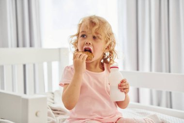 Yatakta oturmak, kurabiye yemek ve süt içmek. Gündüz vakti evde rahat giyimli şirin küçük bir kız var..