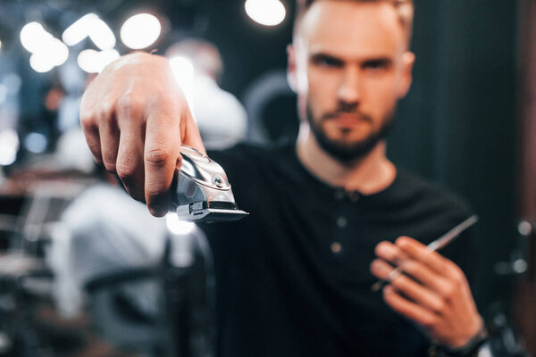 Young bearded man standing in barber shop and holding clipper.