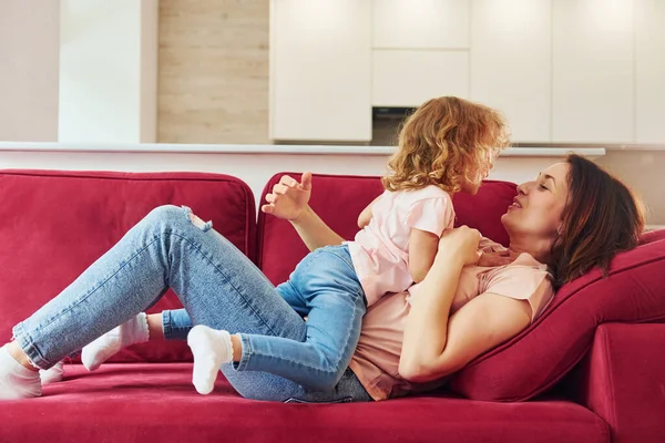 Lying down on red sofa. Young mother with her little daughter in casual clothes together indoors at home.