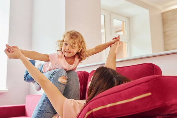 Having fun on red sofa. Young mother with her little daughter in casual clothes together indoors at home.