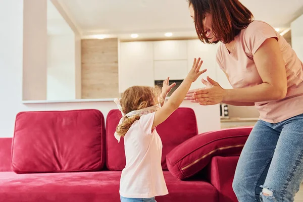 Playing game with eyes blindfolded. Young mother with her little daughter in casual clothes together indoors at home.