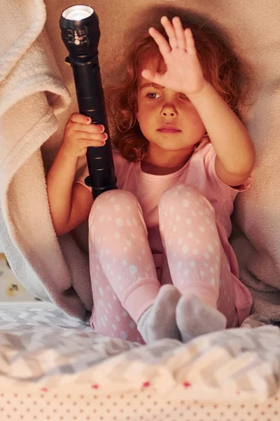 El feneriyle battaniyenin altında oturmak. Gündüz vakti evde rahat giyimli şirin küçük bir kız var..