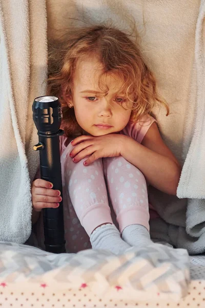 El feneriyle battaniyenin altında oturmak. Gündüz vakti evde rahat giyimli şirin küçük bir kız var..