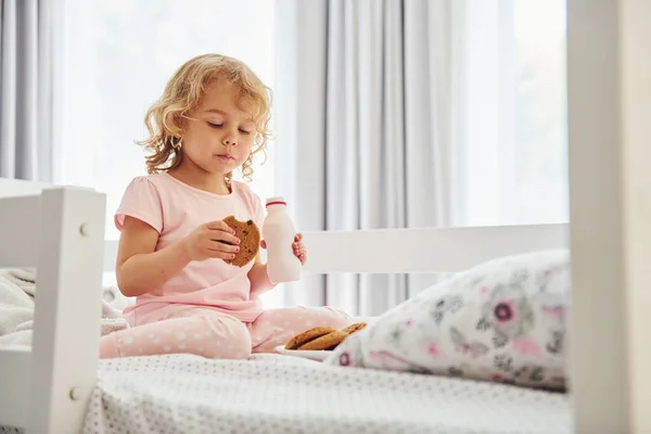 ベッドに座り クッキーを食べ 牛乳を飲む カジュアルな服のかわいい女の子は昼間家にいます — ストック写真