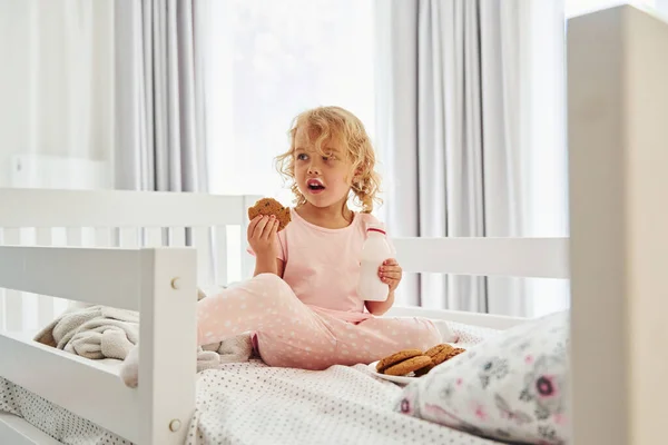 ベッドに座り クッキーを食べ 牛乳を飲む カジュアルな服のかわいい女の子は昼間家にいます — ストック写真