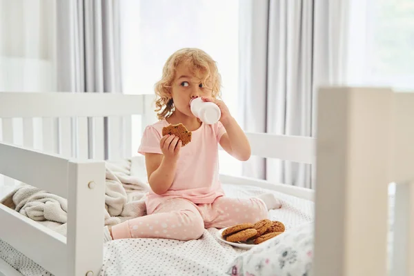 ベッドに座り クッキーを食べ 牛乳を飲む カジュアルな服のかわいい女の子は昼間家にいます — ストック写真