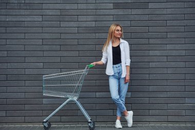 Beautiful blonde in casual clothes with shopping cart is outdoors at sunny daytime.