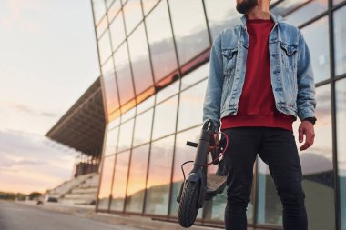 Gündüz vakti iş yerinin yakınında elektrikli ayakkabıyla dışarıda dikilen gündelik kıyafetli yakışıklı bir genç..