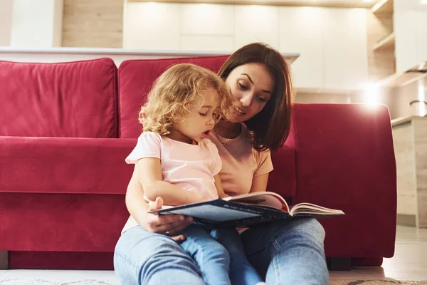 Reading big book. Young mother with her little daughter in casual clothes together indoors at home.