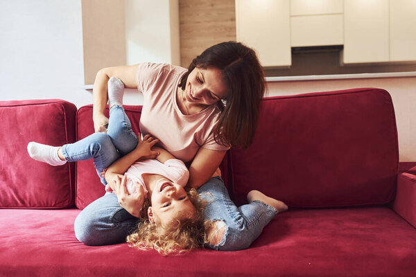Lying down on red sofa. Young mother with her little daughter in casual clothes together indoors at home.