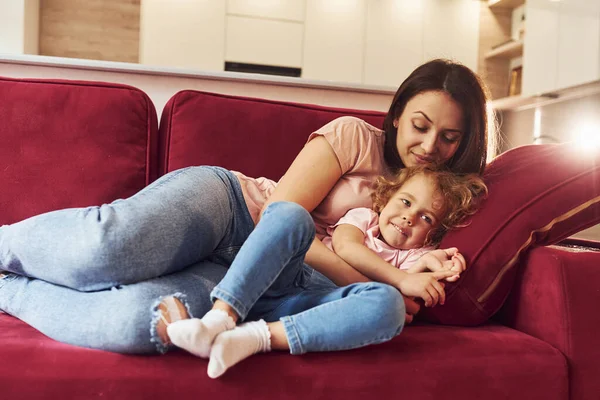 赤いソファの上に横になって 若いです母とともに彼女の小さな娘でカジュアル服で一緒に室内で — ストック写真
