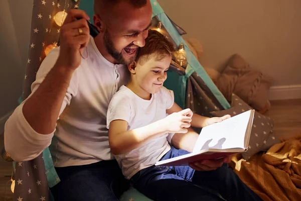 读书和使用手电筒 父亲和他年幼的儿子在帐篷边的屋里呆着 — 图库照片