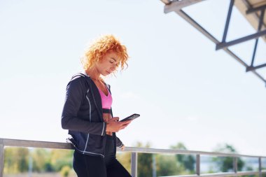 Avrupalı genç, kızıl saçlı, spor elbiseli, açık havada elinde telefonla bekleyen bir kadın..