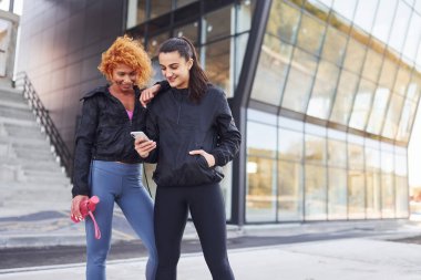 Telefonu elinde tutuyor. Spor vücutlu iki bayan arkadaşın dışarıda spor günü var..
