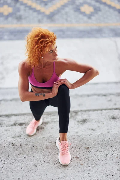 Avrupalı genç kızıl saçlı spor elbiseli kadın dışarıda çömelme yapıyor..