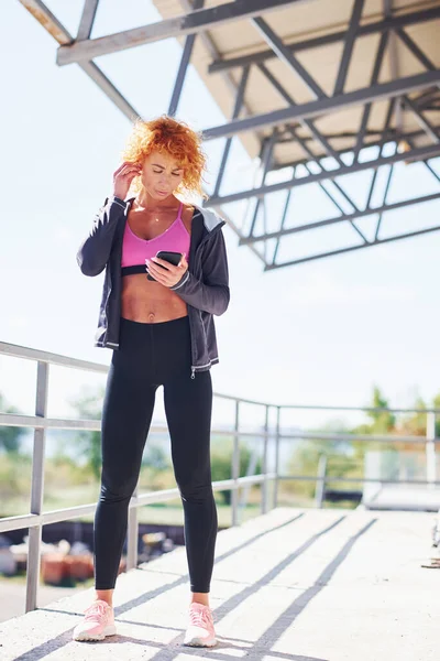 Avrupalı genç, kızıl saçlı, spor elbiseli, açık havada elinde telefonla bekleyen bir kadın..