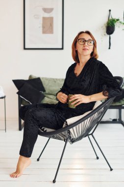 Gözlüklü ve siyah pijamalı hamile bir kadın gündüz vakti evde oturuyor..