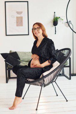 Gözlüklü ve siyah pijamalı hamile bir kadın gündüz vakti evde oturuyor..