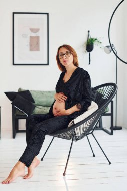 Gözlüklü ve siyah pijamalı hamile bir kadın gündüz vakti evde oturuyor..