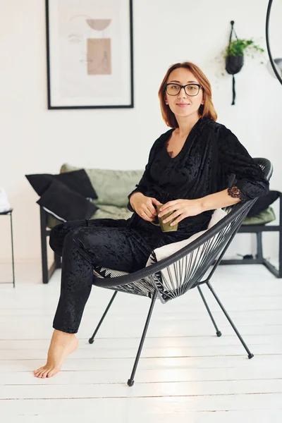 Gözlüklü ve siyah pijamalı hamile bir kadın gündüz vakti evde oturuyor..