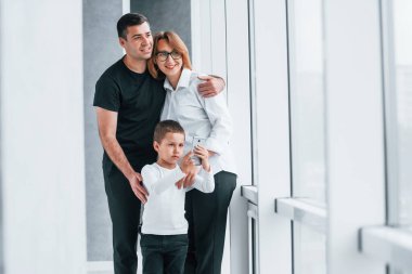 Resmi giyinmiş bir kadın ve elinde kamerayla boş bir odada bir adam ve küçük bir çocukla duruyor..