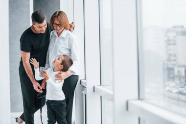 Resmi giyinmiş bir kadın ve elinde kamerayla boş bir odada bir adam ve küçük bir çocukla duruyor..