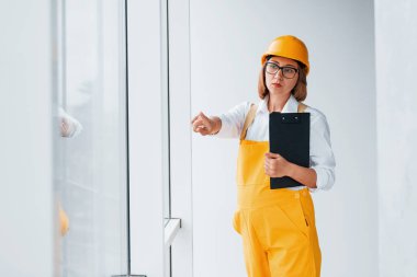 Elinde not defteriyle. Sarı üniformalı kadın işçi ya da mühendis ve içeride duran sert şapkalı..