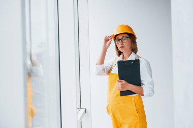 Elinde not defteriyle. Sarı üniformalı kadın işçi ya da mühendis ve içeride duran sert şapkalı..