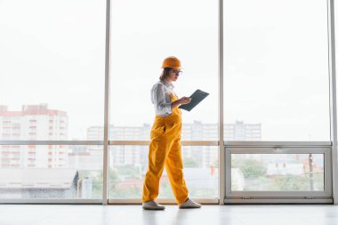 Büyük pencerenin yanında. Sarı üniformalı kadın işçi ya da mühendis ve içeride duran sert şapkalı..